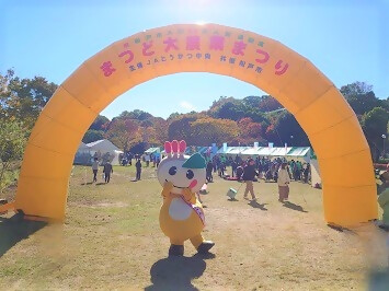 まつど大農業まつりのアーチでポーズを決めるかまたんの写真